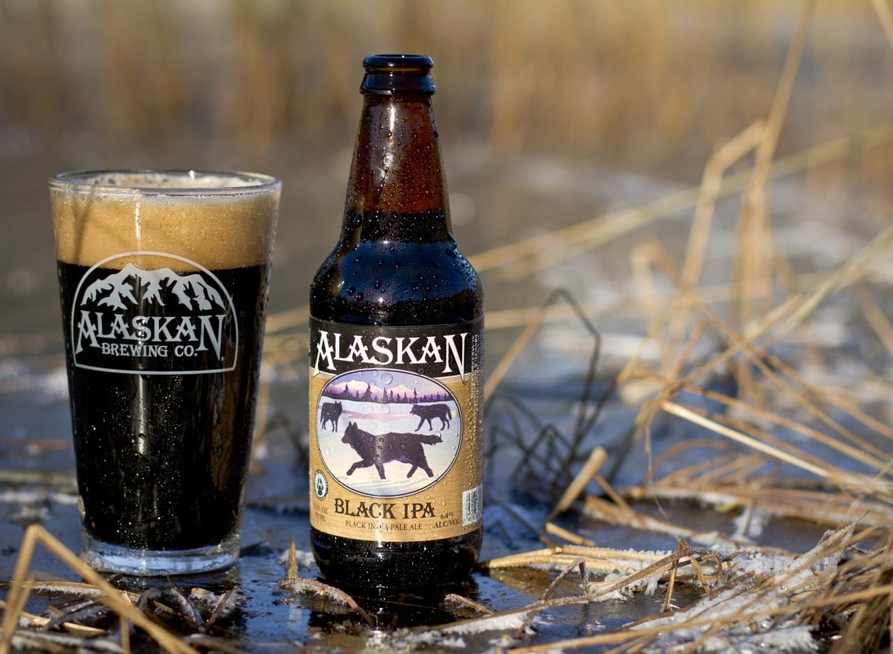 Alaskan Brewing Co. Black IPA bottle and glass on snow-covered ground with grass blades.