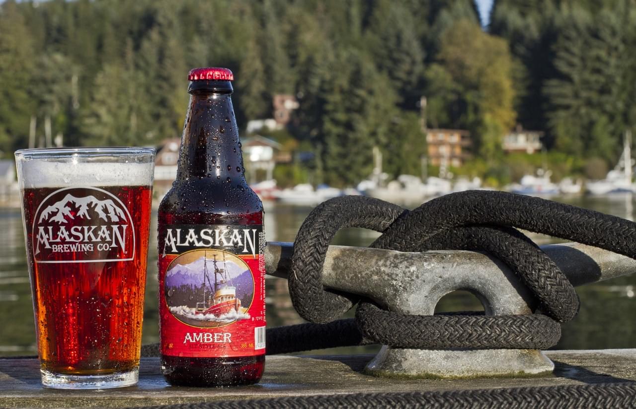 Alaskan brewing beer bottle and glass on dock near water, outdoor scene with boats and forest backdrop.
