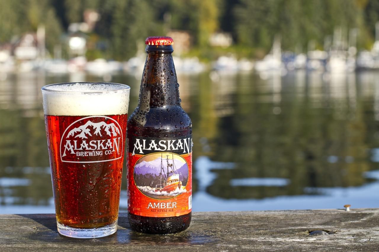 Alaskan Amber beer bottle and glass on a wooden dock, with a scenic water background and boat, outdoor leisure drink.