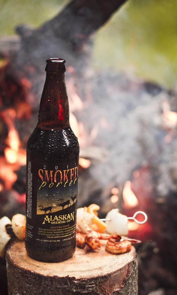 Dark beer bottle with smoked flavors, Alaskan Brewing Co., outdoor campfire scene.