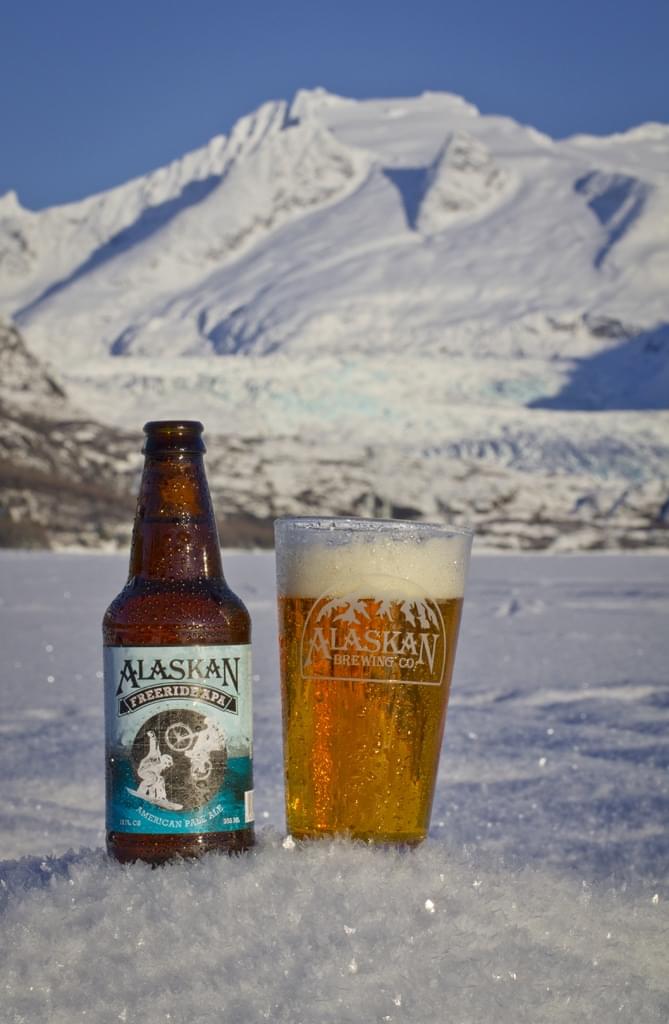 Alaskan Brewing Company beer bottle and glass in snowy mountain landscape, cold climate, outdoor pub, refreshing summer drink.