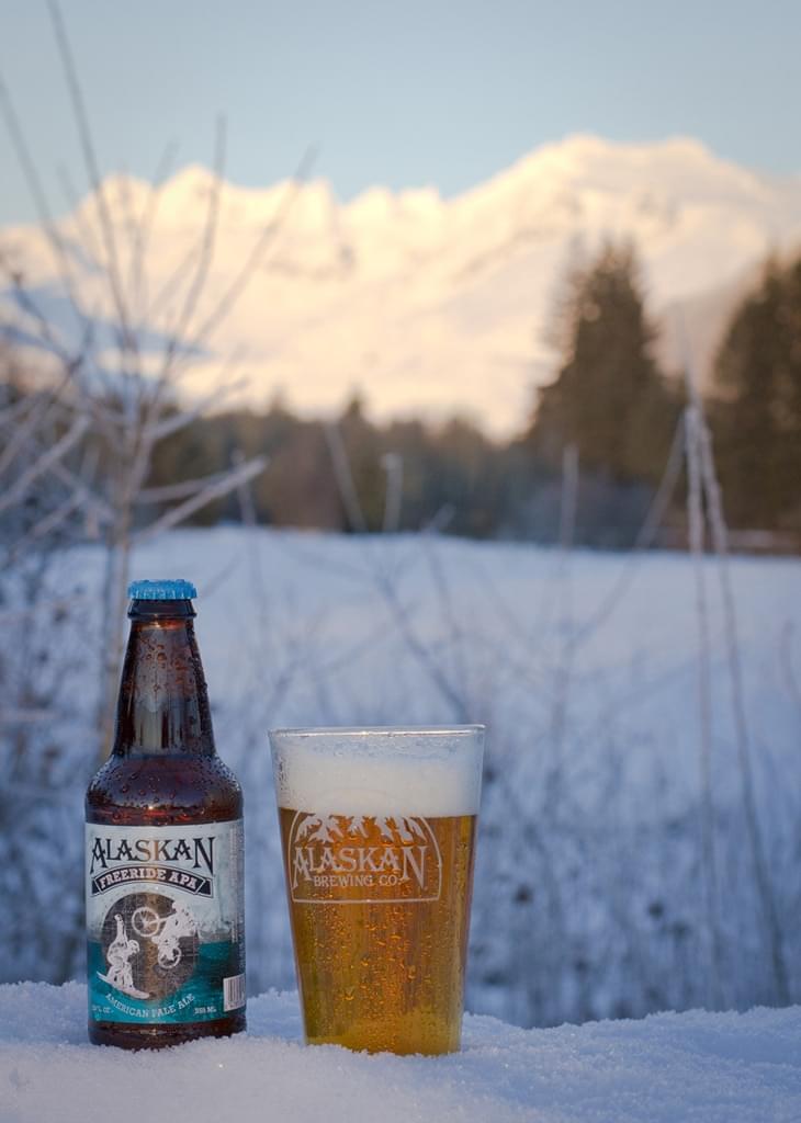 Alaskan Brewing beer bottle and glass in a snowy landscape with mountain views.