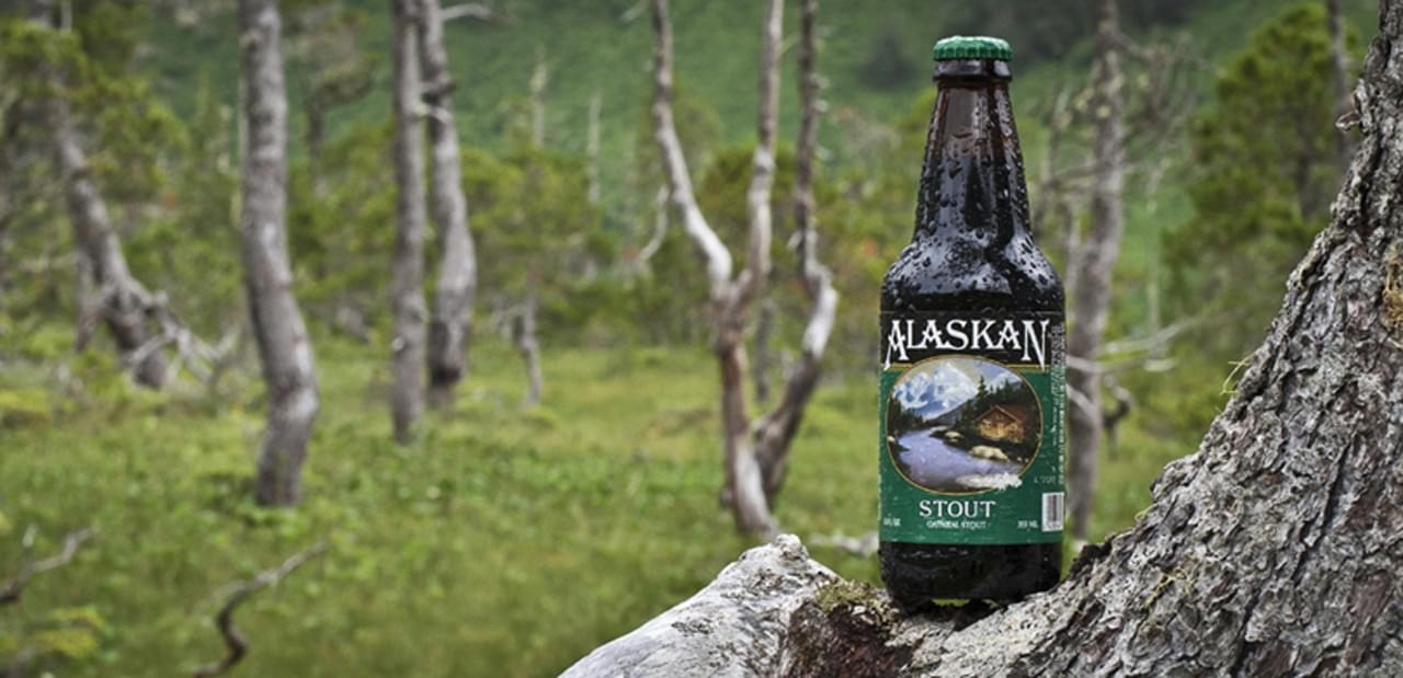 Alaskan Stout beer bottle outdoors in nature forest setting, featuring mountain and river scenery on label.